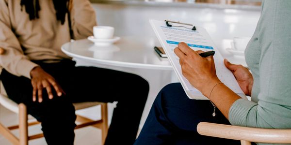 Two people sitting at a table. One with a cup of tea, the other with a clipboard.