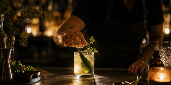 Bartender garnishing a cocktail with a green leaf in a dimly lit bar.