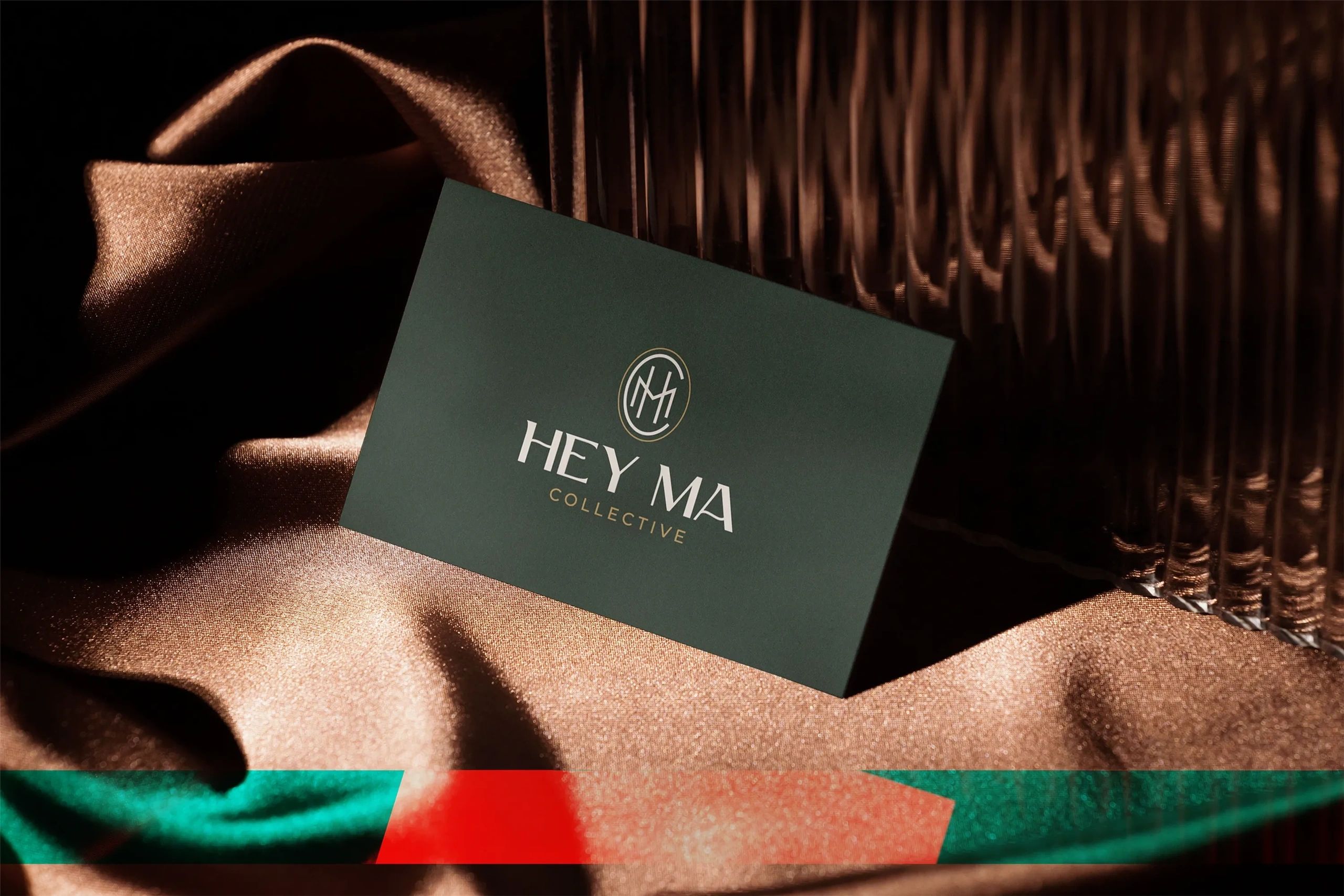 Dark green Hey Ma Collective business card with cream and gold logo displayed on bronze fabric.