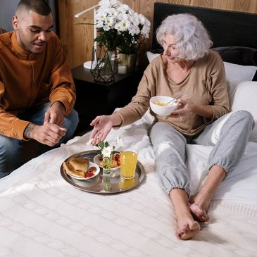 A young man seated by the bedside of an elderly lady having breakfast.
