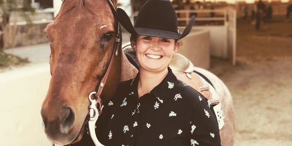 Smiling woman in cowboy attire standing next to a saddled horse.