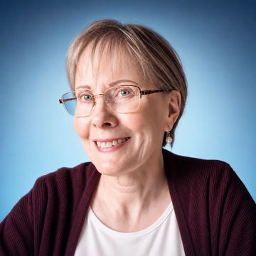 Smiling elderly woman wearing glasses and a maroon cardigan against a blue background.