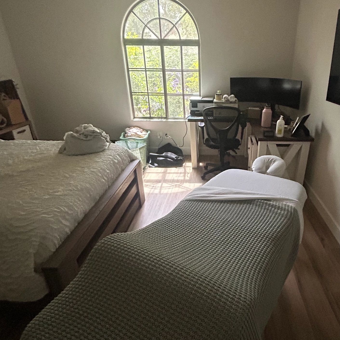 Cozy room with a massage table, bed, desk, and arched window letting in natural light.