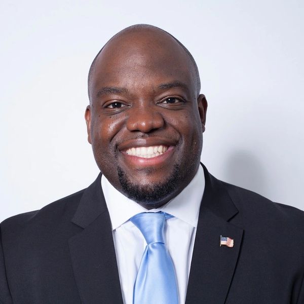 Smiling man in suit with light blue tie and American flag pin.