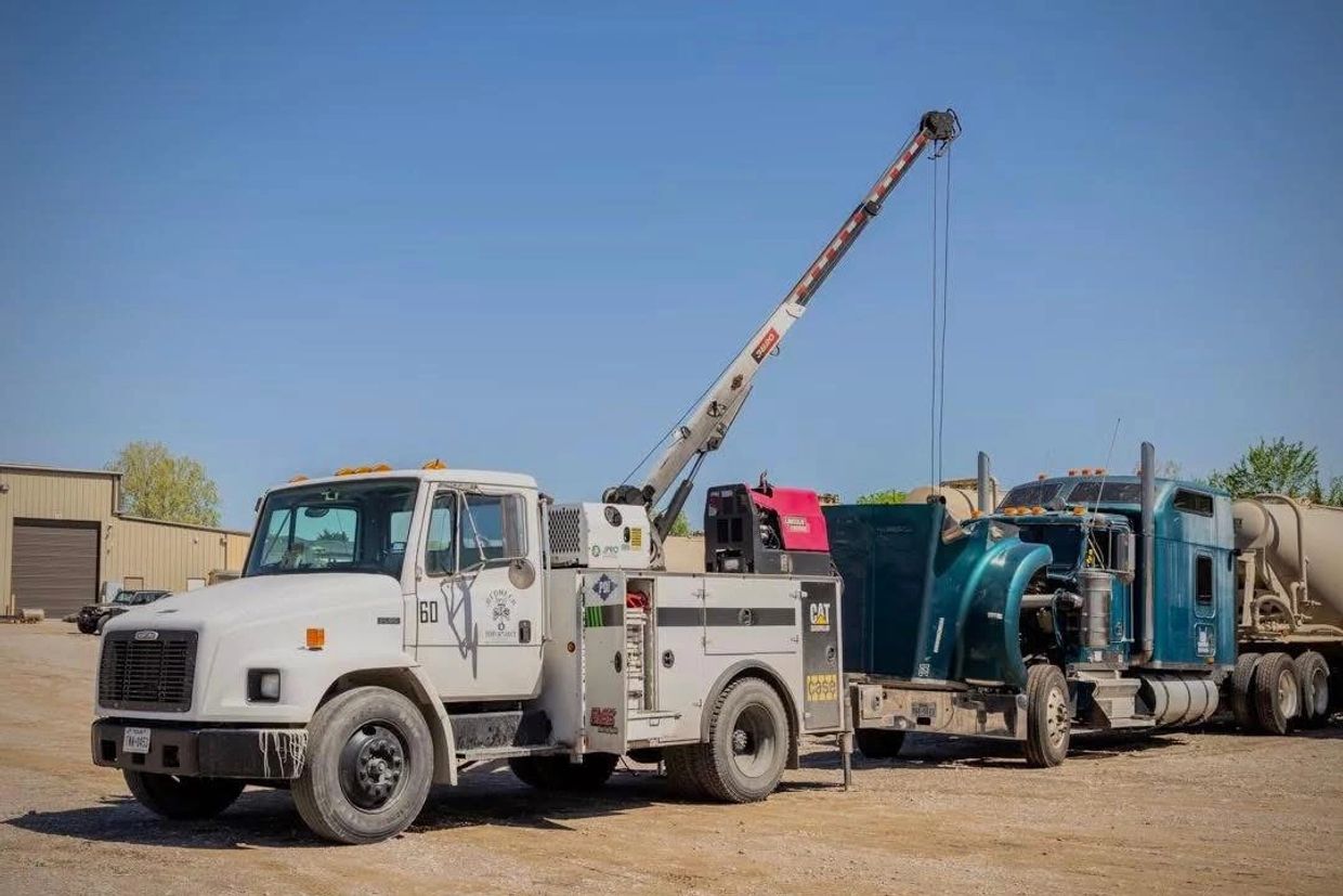 A white tow truck lifting a blue semi-truck's engine hood in a sunny yard.