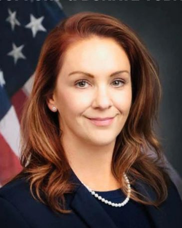 Professional woman with red hair and pearl necklace in front of American flag.