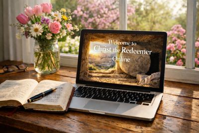 Laptop on wooden desk showing a religious welcome message with flowers and a Bible nearby.