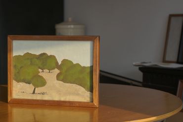 A framed landscape painting of trees on a table in a dim room.