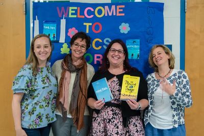 Four women in front of a bulletin board at school holding books