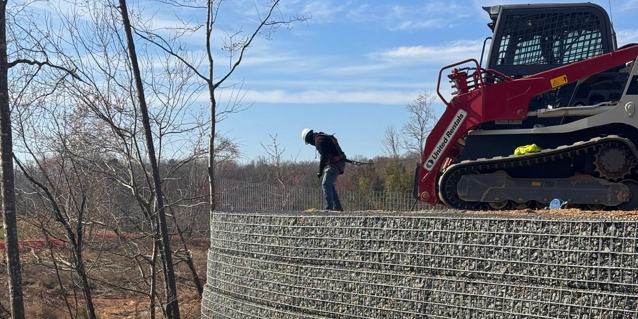 Construction worker secured with harness on a stone wall near heavy machinery.