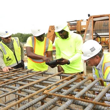 Construction workers reviewing plans on a site with rebar and heavy machinery.