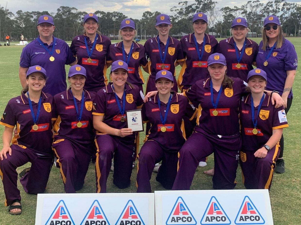 A victorious women's cricket team posing with medals and a plaque on the field.