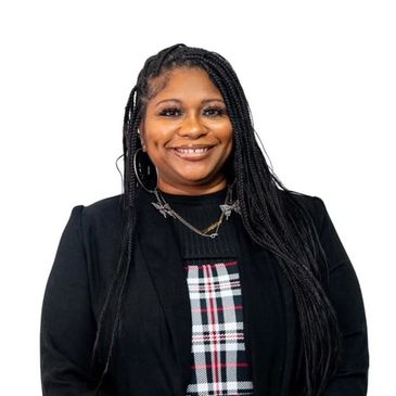Smiling woman with long braided hair wearing a black blazer and plaid dress.