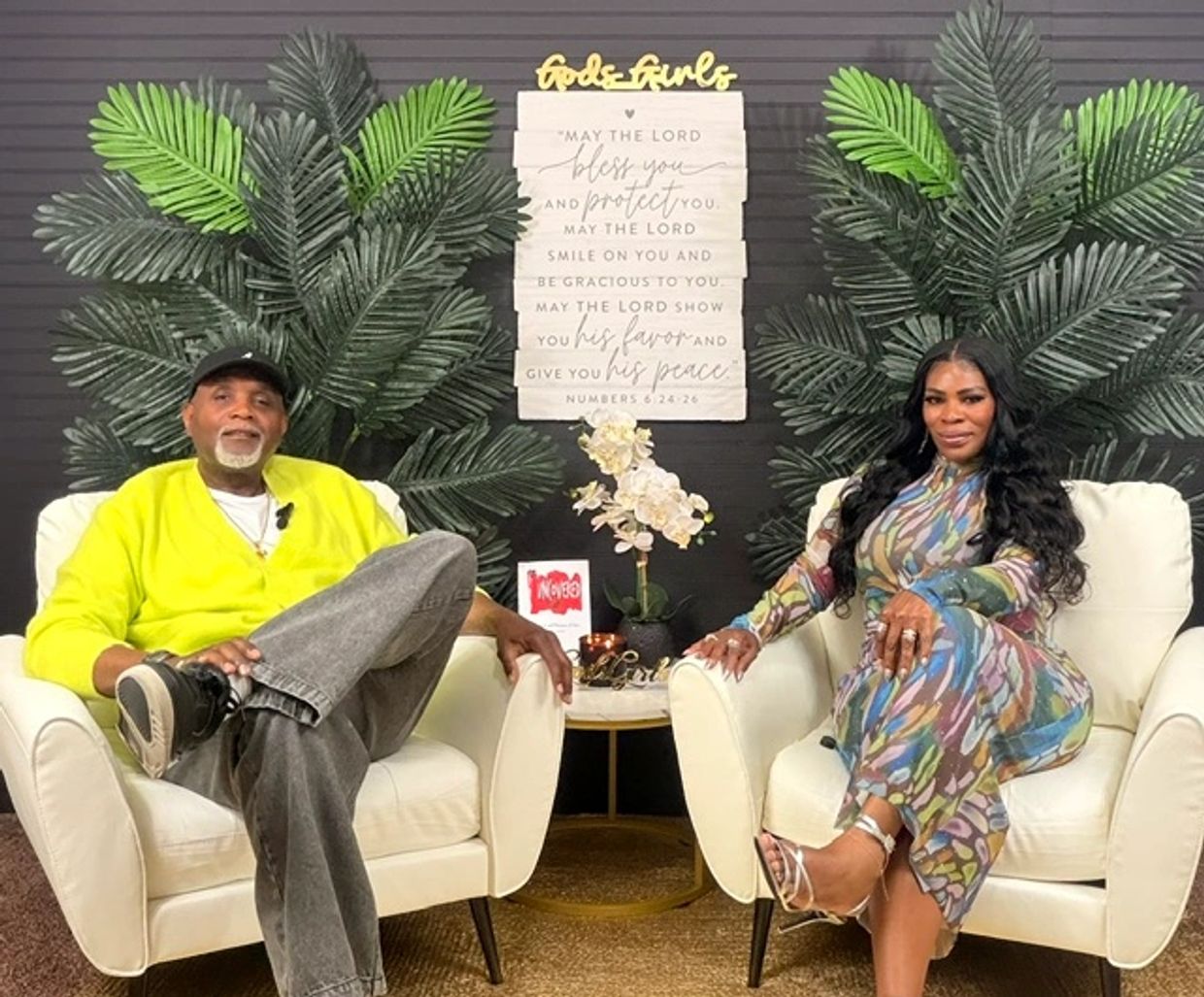 Two people seated in white chairs with green plants and inspirational decor behind them.