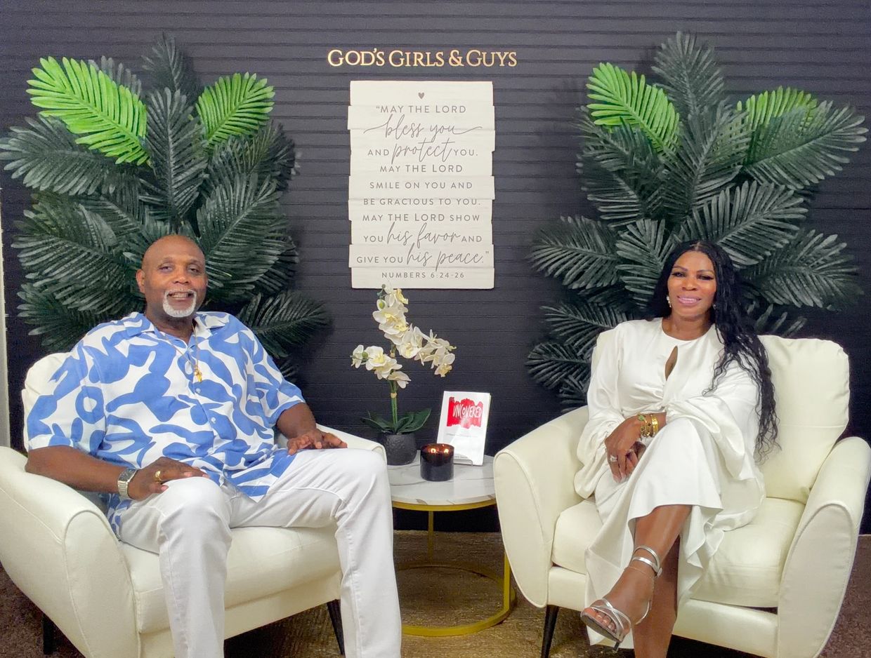 A man and woman seated in white armchairs with a religious wall sign behind.