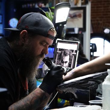Tattoo artist focused on inking a client's arm with a Joker image reference.