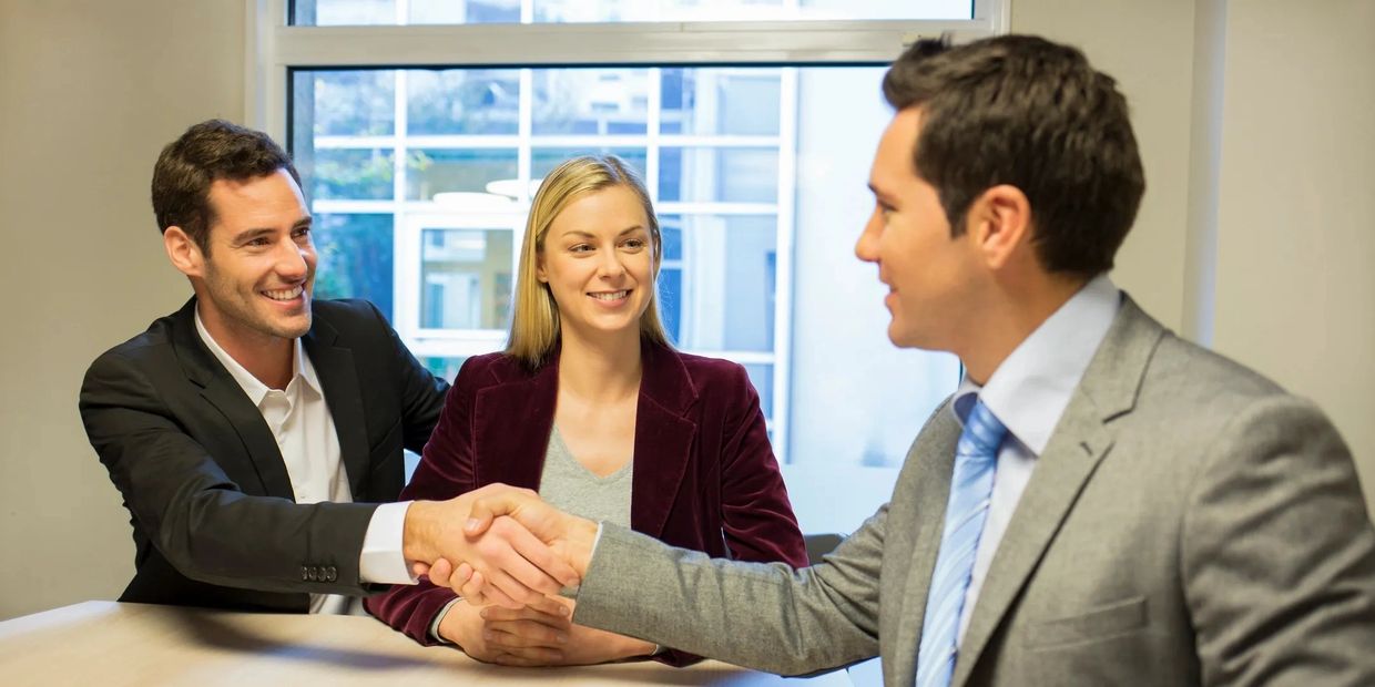 Two men shaking hands with a woman smiling between them in a bright office.