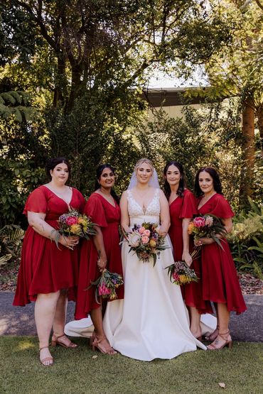 Bride in white gown with bridesmaids in red dresses holding bouquets outdoors.