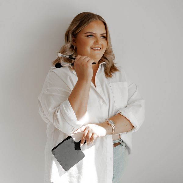 Smiling woman holding makeup brushes and a mirror, dressed in a white shirt.