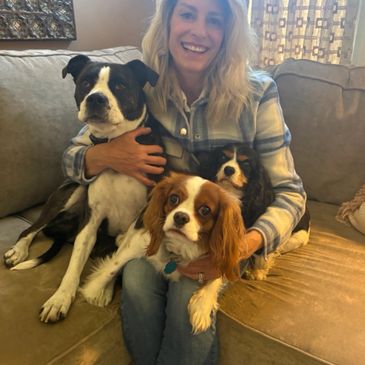 A woman sits on a couch smiling with three dogs in her lap.