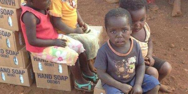 Children sitting on boxes of hand soap outdoors on dirt ground.