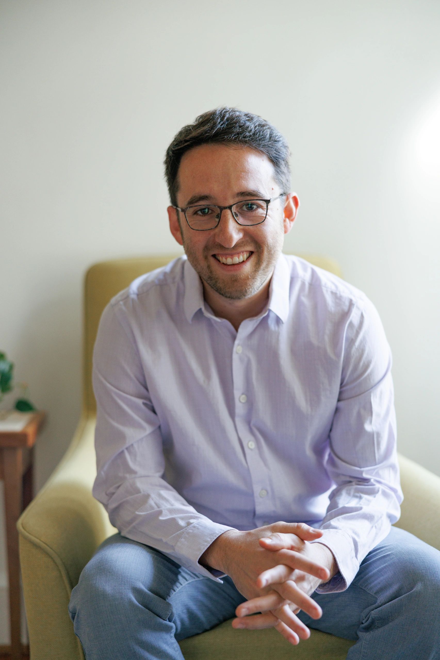 Sito is seated and smiles warmly at the camera in a white button-up shirt, jeans, and glasses 