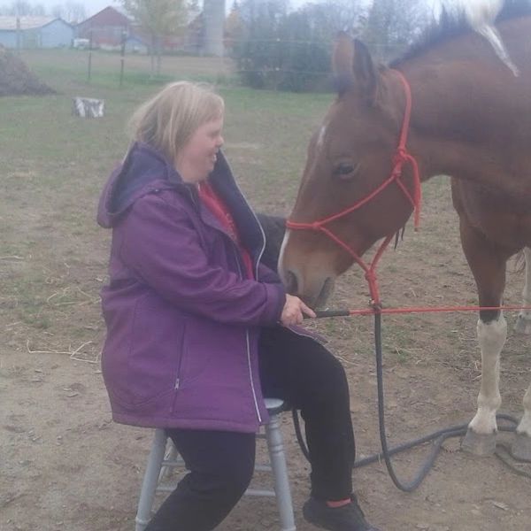 A woman in a purple jacket sitting on a stool, interacting with a brown and white horse.