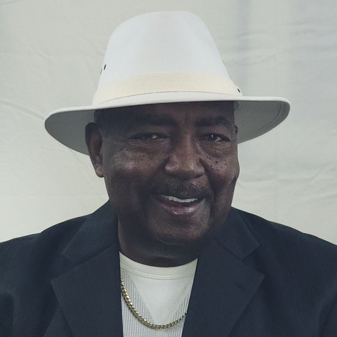 Smiling man wearing a white hat, gold chain, black jacket, and white shirt.