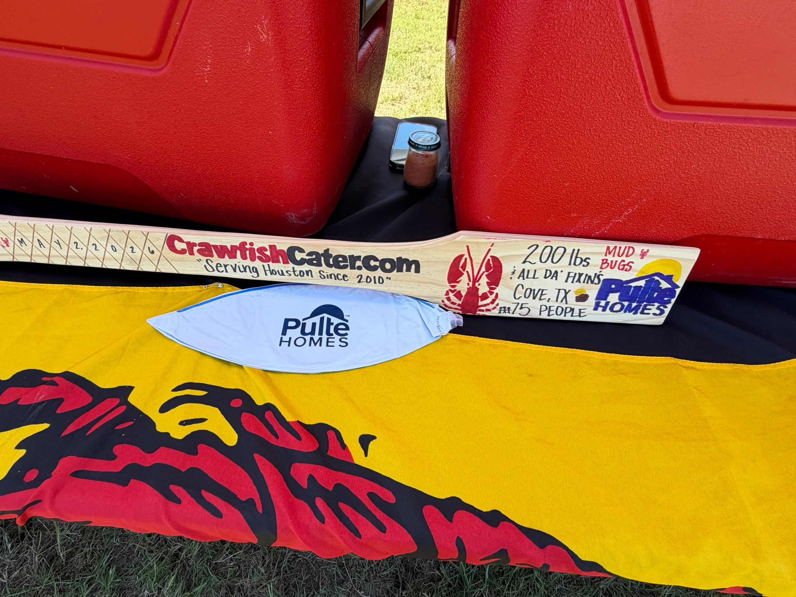 Custom wooden crawfish paddle with sponsor logos between red coolers.