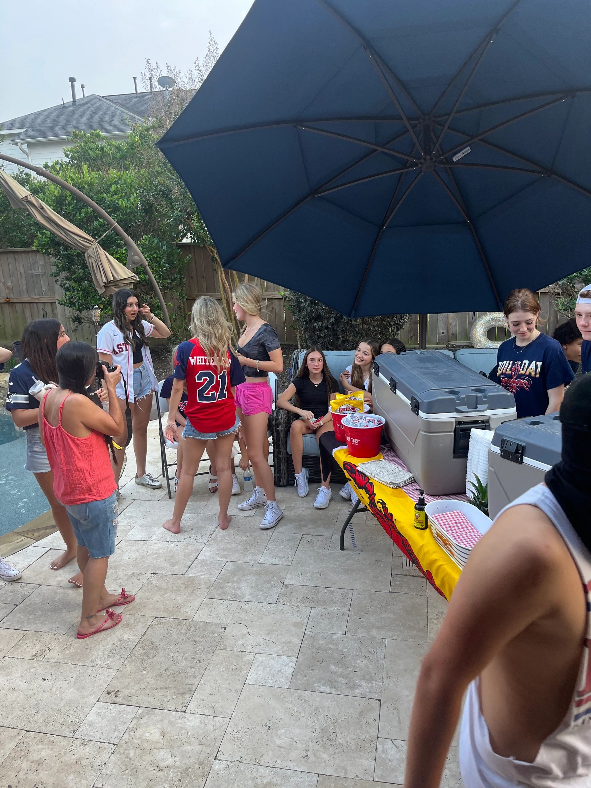 Group of young people socializing outdoors near a pool with large blue umbrella.