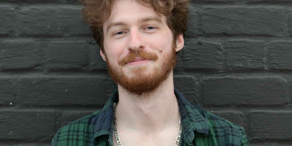 Smiling young man with curly hair and beard in front of a dark brick wall.