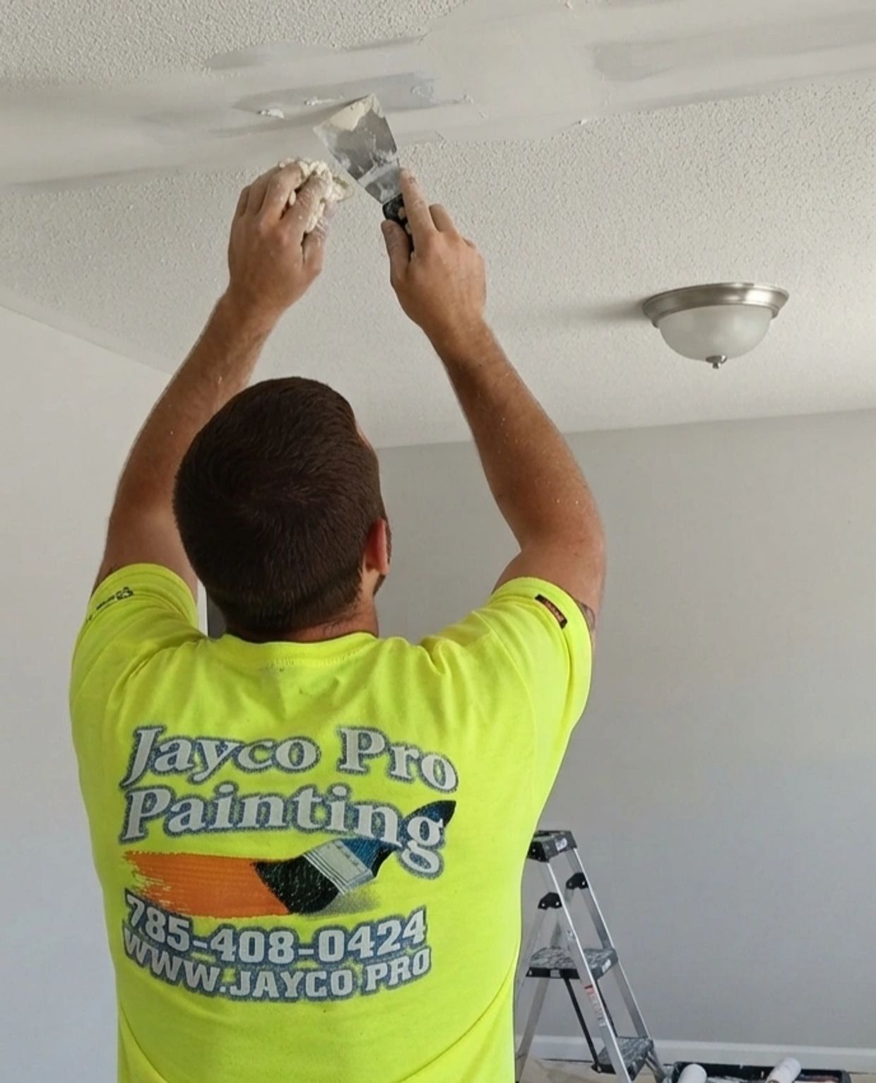 A painter wearing a JaycoPro  Painting work shirt is removing popcorn texture from a ceiling. 