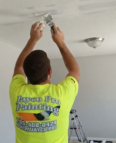 A painter wearing a Jayco apron Painting work shirt is removing popcorn texture from a ceiling. 