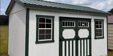 White storage shed with green trim and dual windows under a partly cloudy sky.