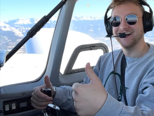 A pilot smiling and giving thumbs up inside a cockpit with mountains outside.