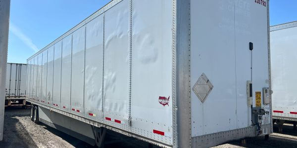 White semi-trailer with dents on its side parked under clear blue sky.