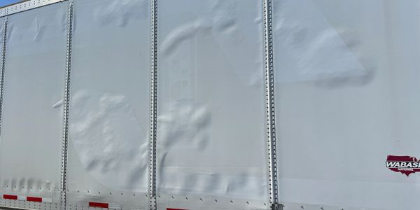 Side view of a white semi-trailer with dents and a Wabash logo.