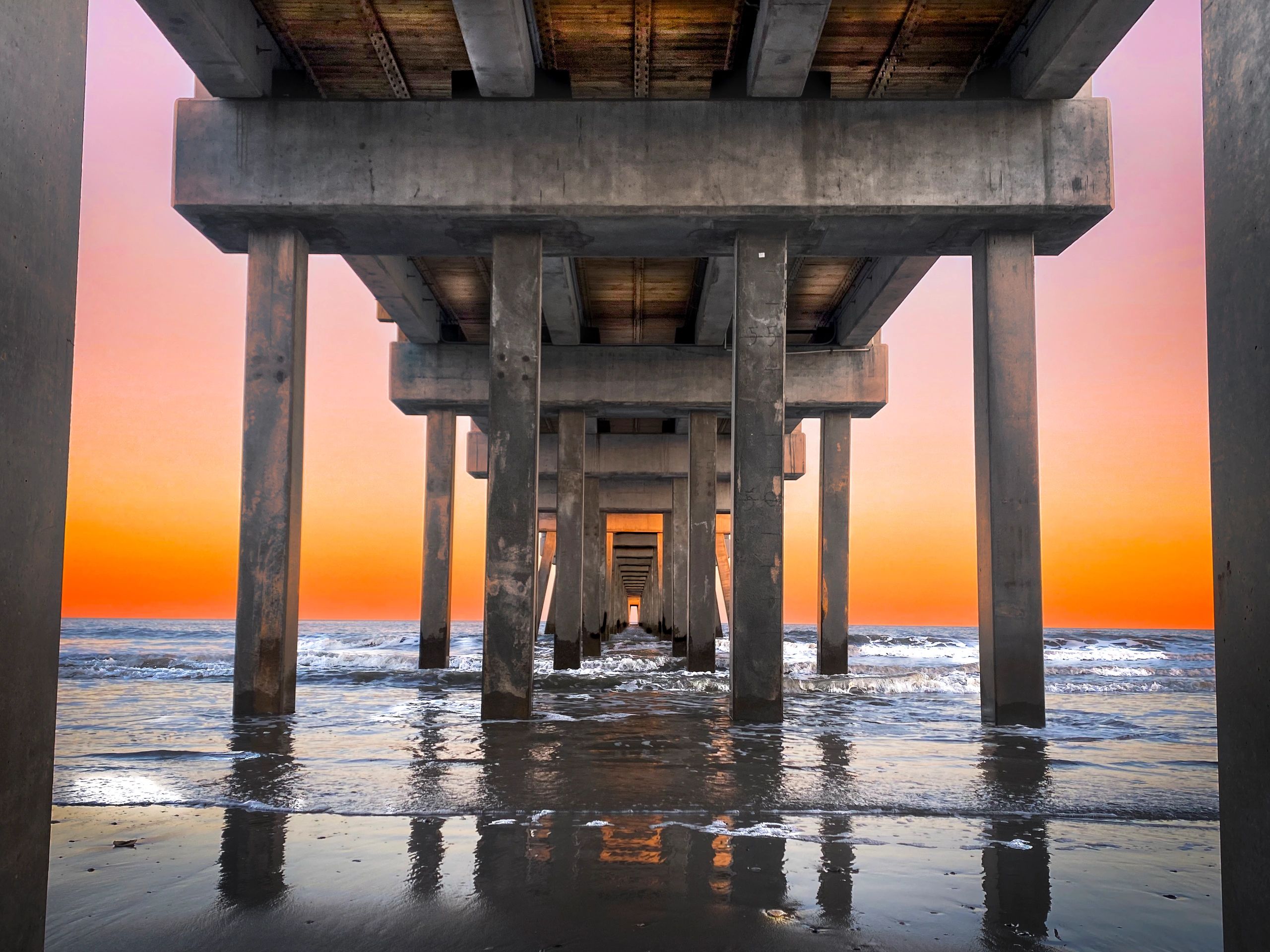 Underneath a pier at sunset with ocean waves.