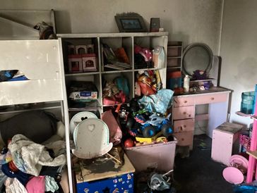 Cluttered children's room with toys, clothes, and furniture scattered around.
