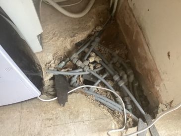 Exposed plumbing pipes in a partially demolished floor corner near a washing machine.