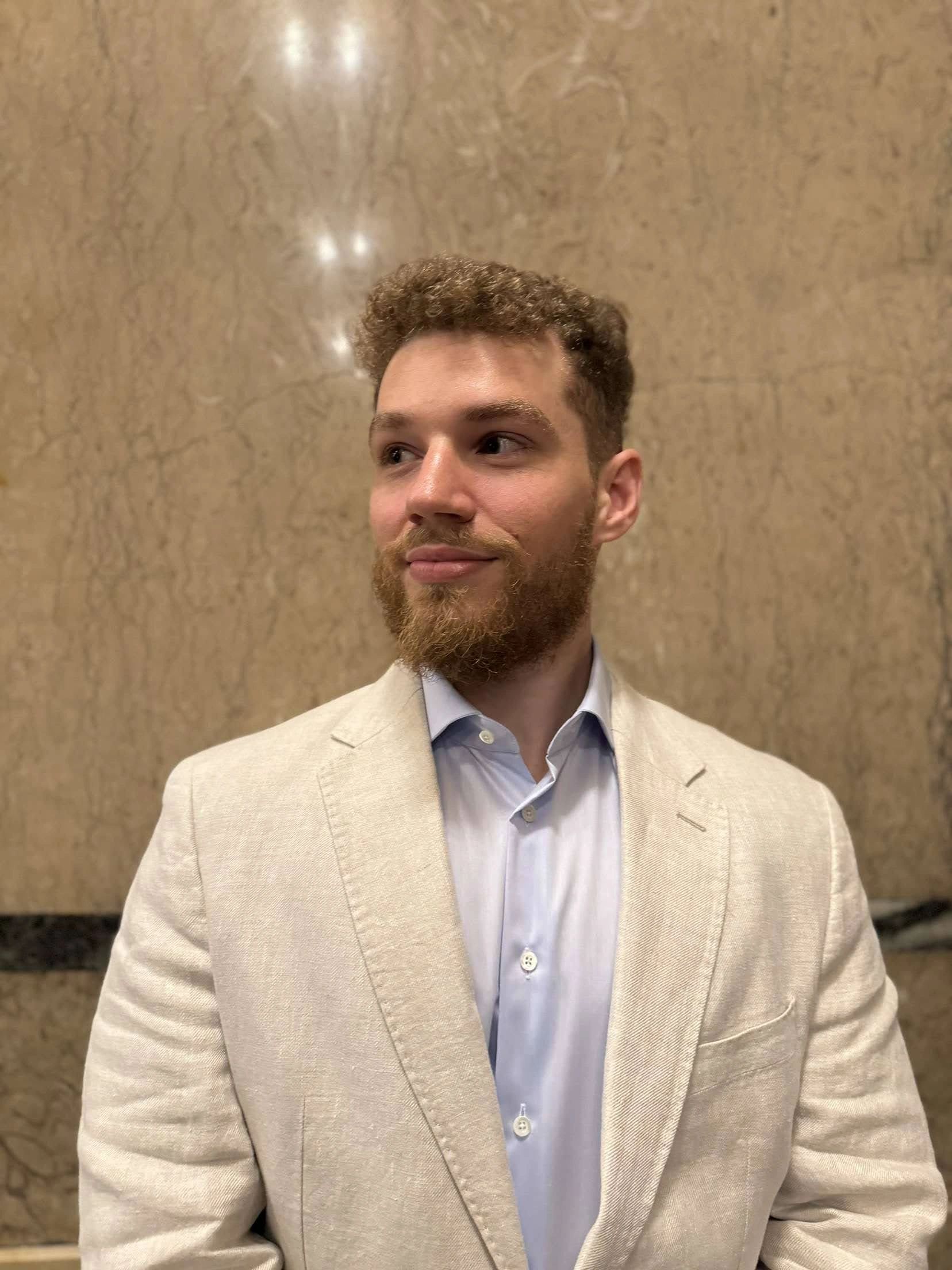 Man in a beige blazer and light blue shirt against a marble wall.