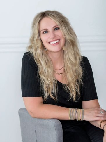 Smiling woman with long blonde hair wearing a black outfit, sitting on a gray chair.