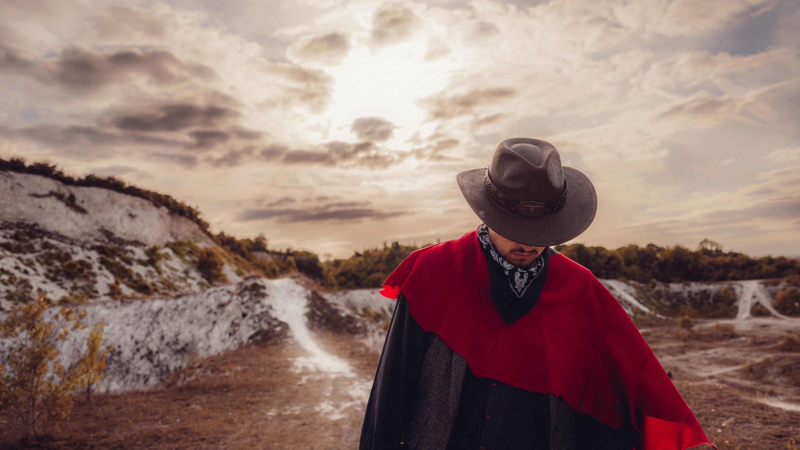 A person in a cowboy hat and red cloak under a cloudy sky.