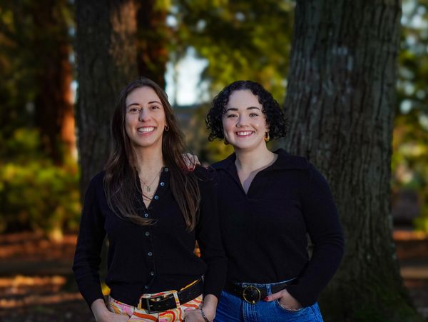 Two women smiling outdoors against a backdrop of trees and sunlight.
