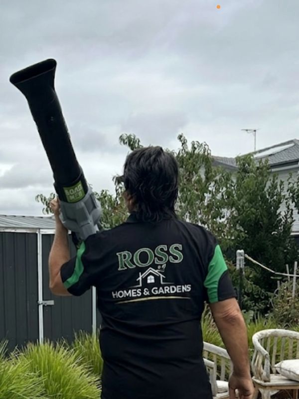 A man holding a leaf blower in a garden wearing a 'ROSS Homes & Gardens' shirt.