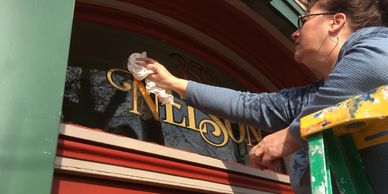 A woman cleans gold lettering on a glass window from a ladder.