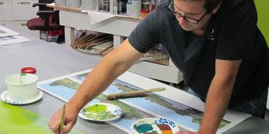 Artist painting a landscape scene on large paper at a studio table.