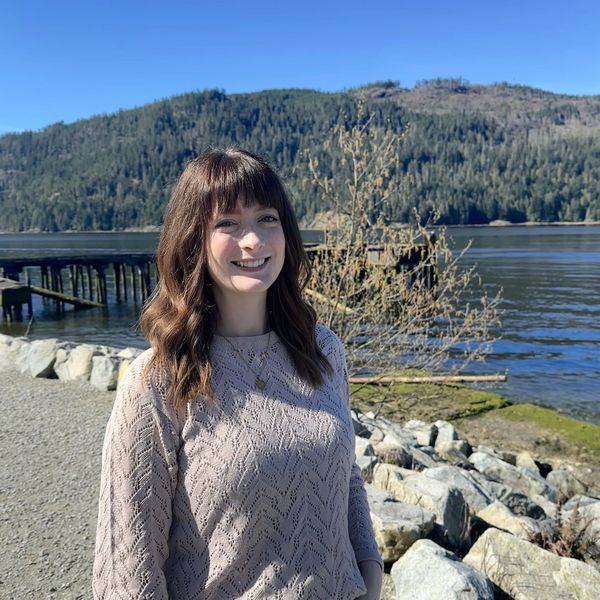 A woman smiling by a lakeside with mountains in the background.