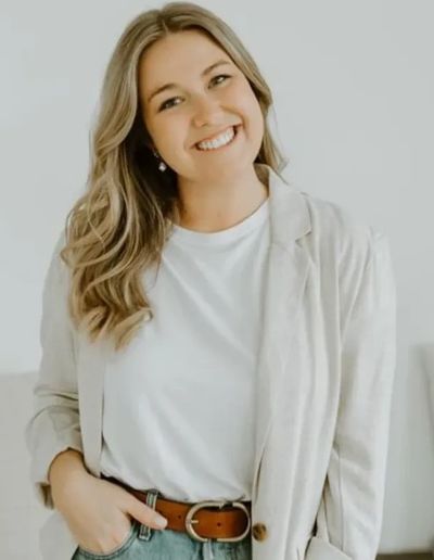 Smiling woman with wavy hair wearing a white shirt and beige jacket.