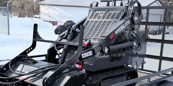 Compact industrial skid steer loader on a trailer in a snowy outdoor area.